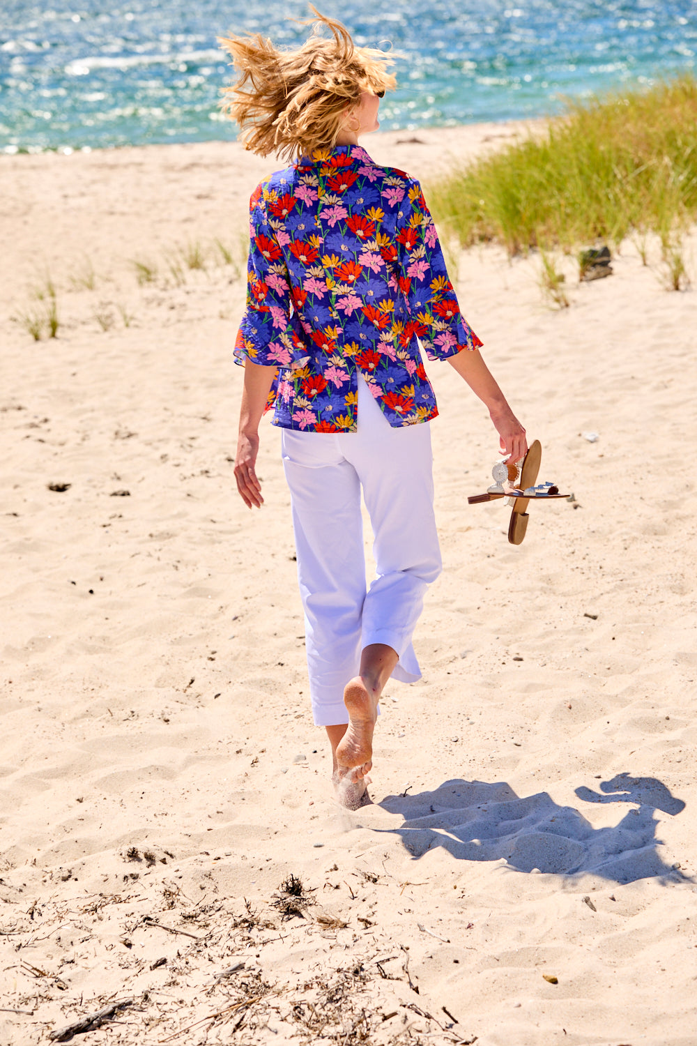 Person walking on a sandy beach wearing a colorful floral shirt and white pants.