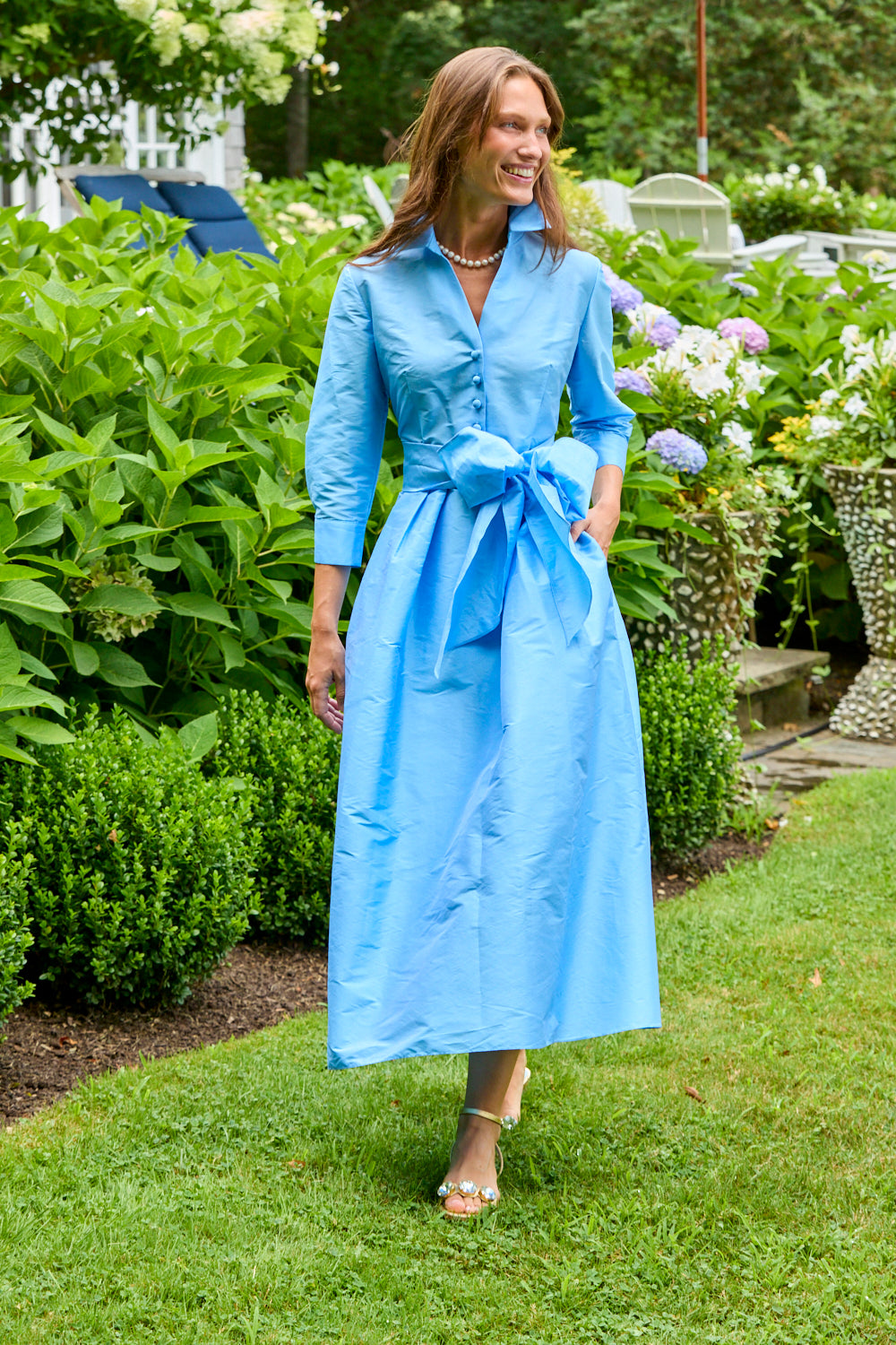 The Ainsley Dress - Light Blue worn by a woman in a garden featuring a long sleeve button front design tie waist and elegant maxi length silhouette
