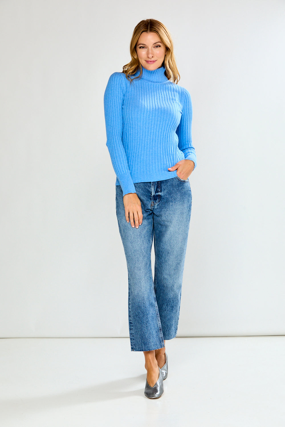 Woman wearing a blue ribbed turtleneck sweater and jeans on a plain background