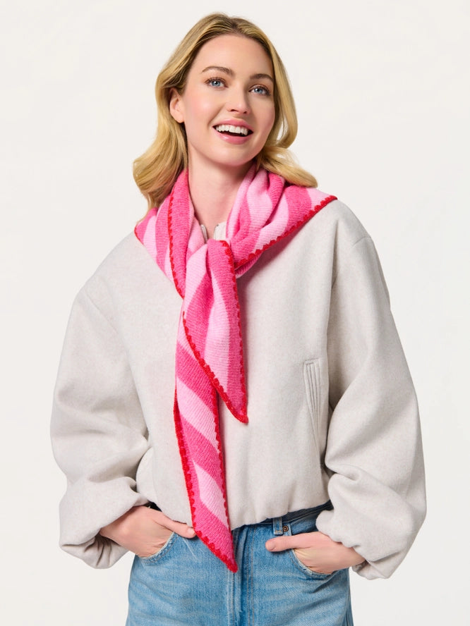 A smiling blonde woman wearing a light gray oversized sweater, blue jeans, and a bright pink striped knit scarf with red fringe, posed against a clean off-white studio background.