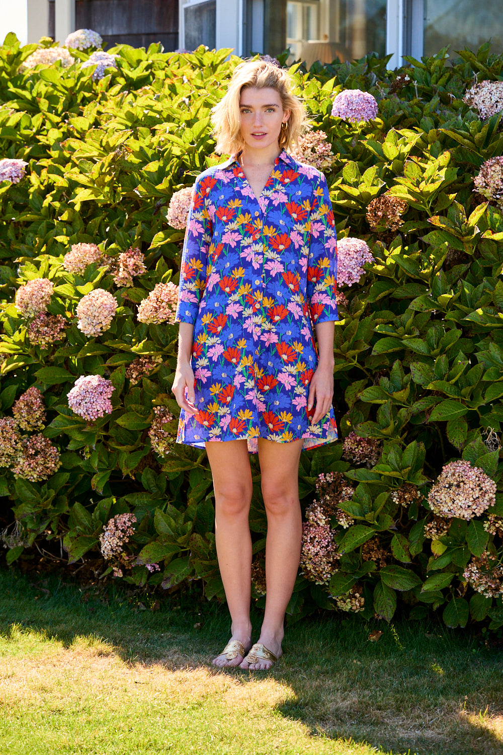 Woman in a colorful floral dress standing in front of a hedge with flowers
