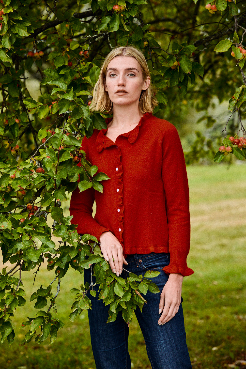 Woman wearing a red shirt and blue jeans standing in a natural setting with greenery.