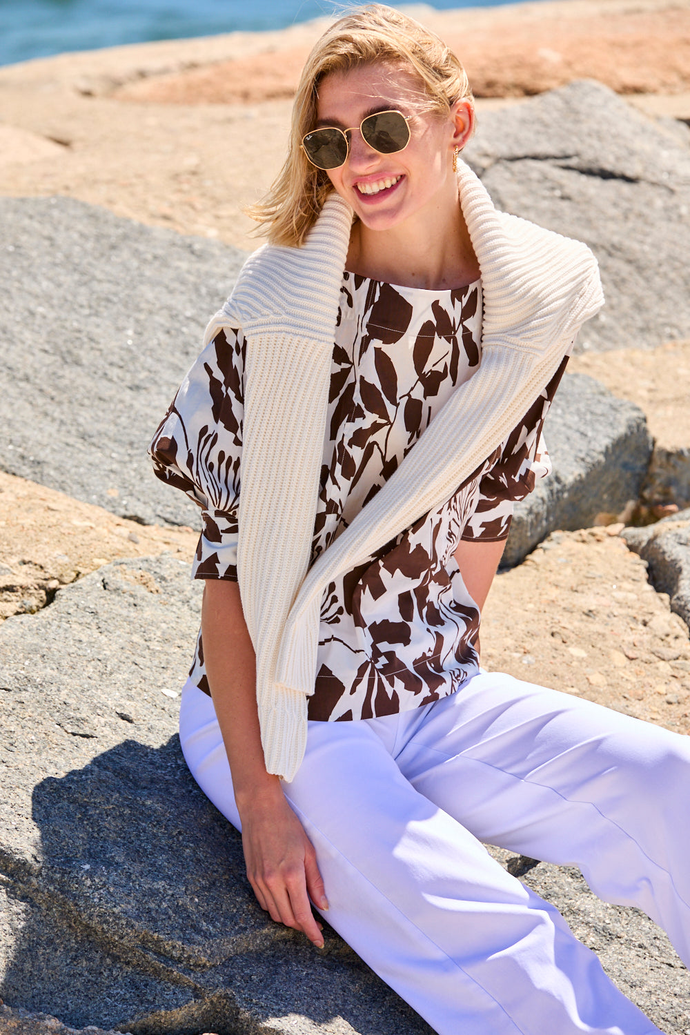 Woman in a patterned top and white pants standing on a rocky beach.