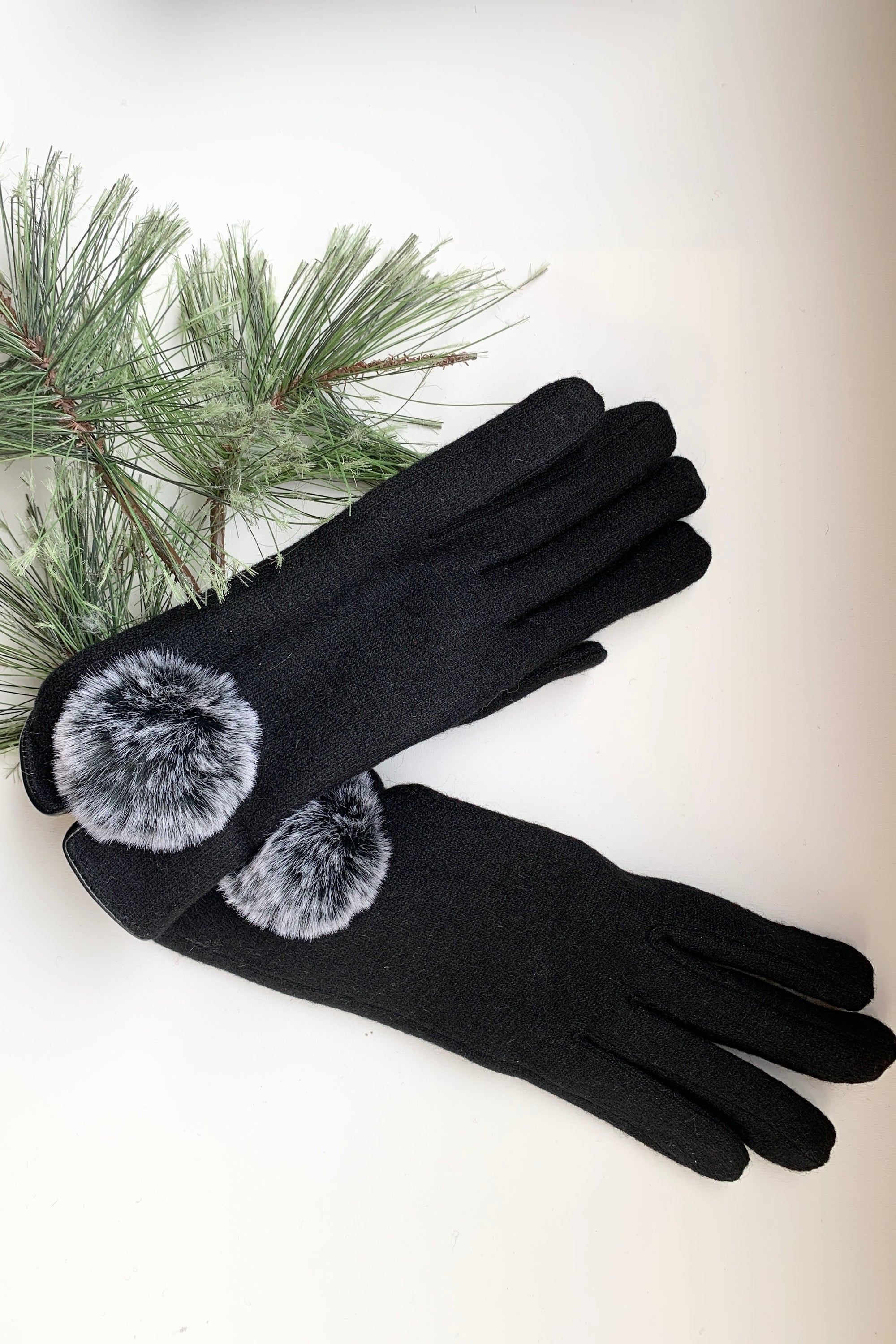 Black gloves with gray pom-poms on a white background with greenery.