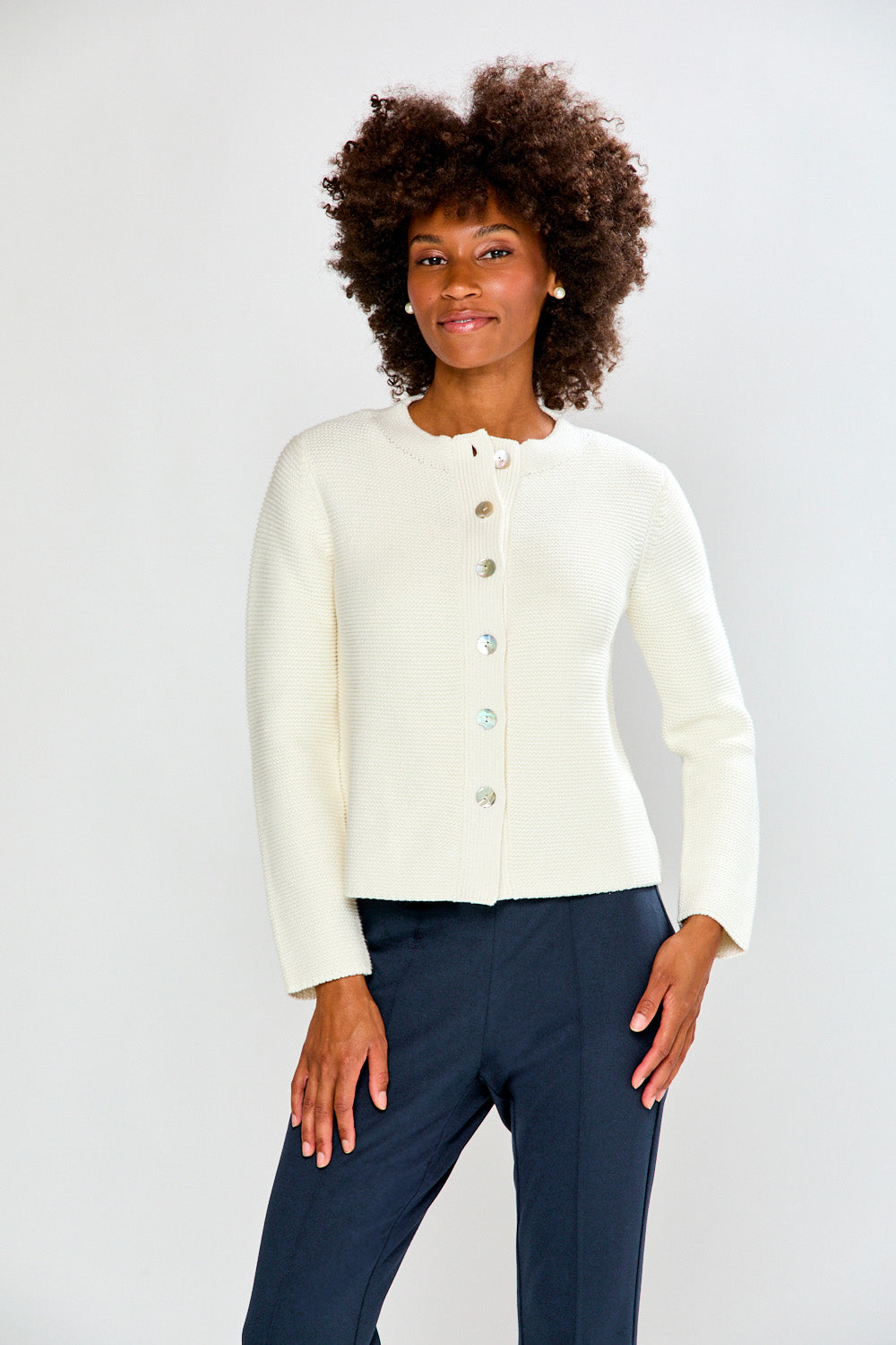 Woman wearing a white cardigan and navy pants against a plain background
