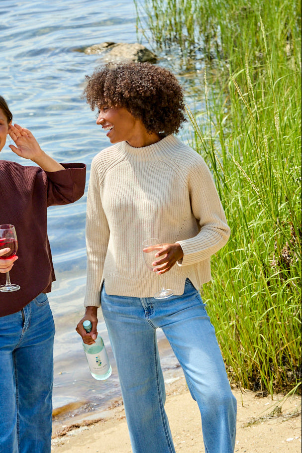 Two women standing on a sandy lakeshore wearing light-wash blue jeans and knit tops, one holding a wine glass and the other holding a bottle, with water and reeds in the background.