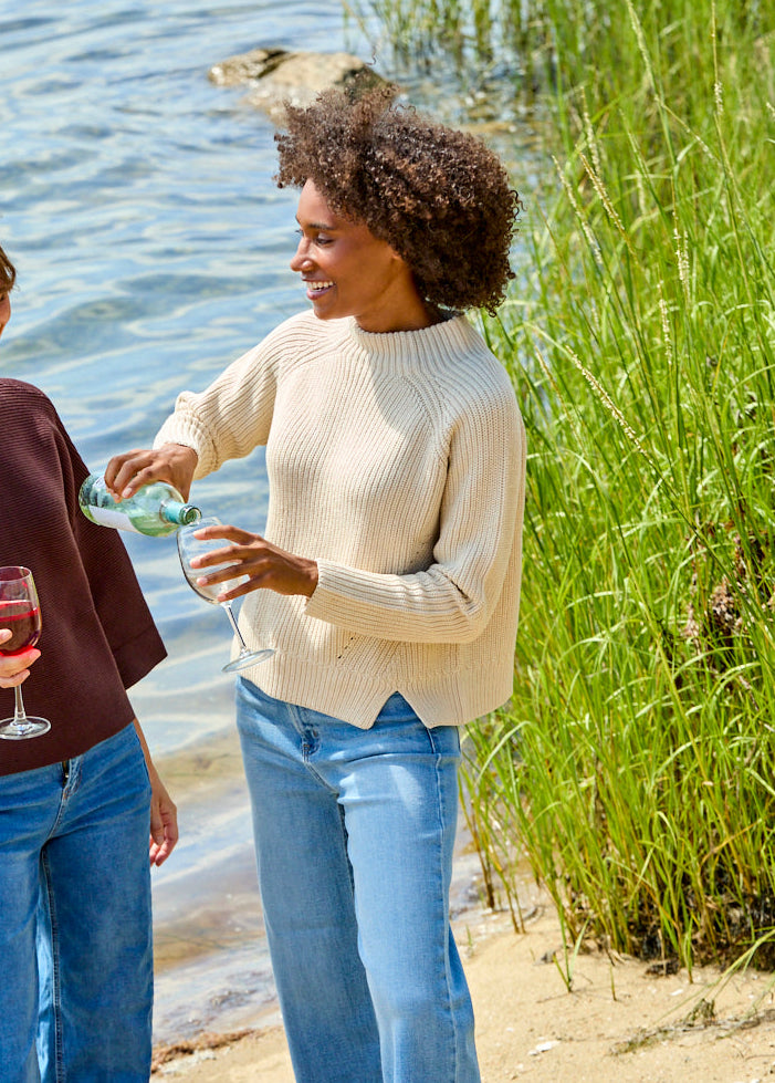 Two women standing on a sandy lakeshore by the water, one holding a wine glass and the other pouring wine, wearing casual knit tops and jeans.