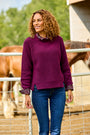 A smiling woman with curly brown hair stands outdoors in front of a wire fence, wearing a ribbed long-sleeve sweater in deep burgundy-purple with patterned lace trim at the collar and cuffs, paired with blue jeans.