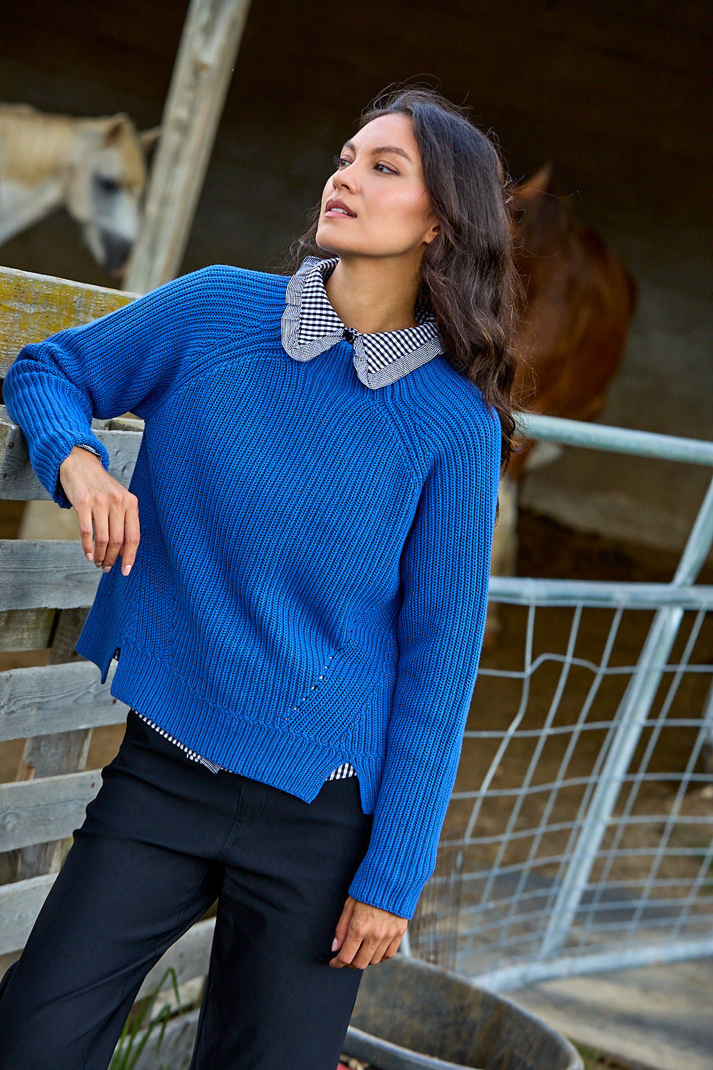 A woman wearing a bright blue ribbed knit sweater with a black and white gingham check collar, paired with black trousers, posing in a rustic barn setting with horses in the background.