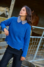 A woman wearing a bright blue ribbed knit sweater with a black and white gingham check collar, paired with black trousers, posing in a rustic barn setting with horses in the background.