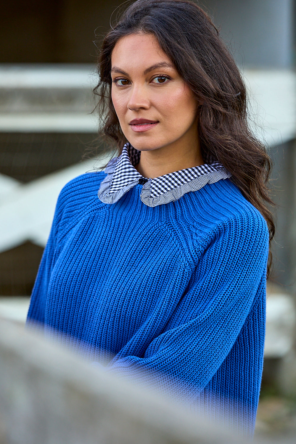 A woman wearing a bright blue ribbed knit sweater with a black and white checkered collar, photographed outdoors against a blurred background.