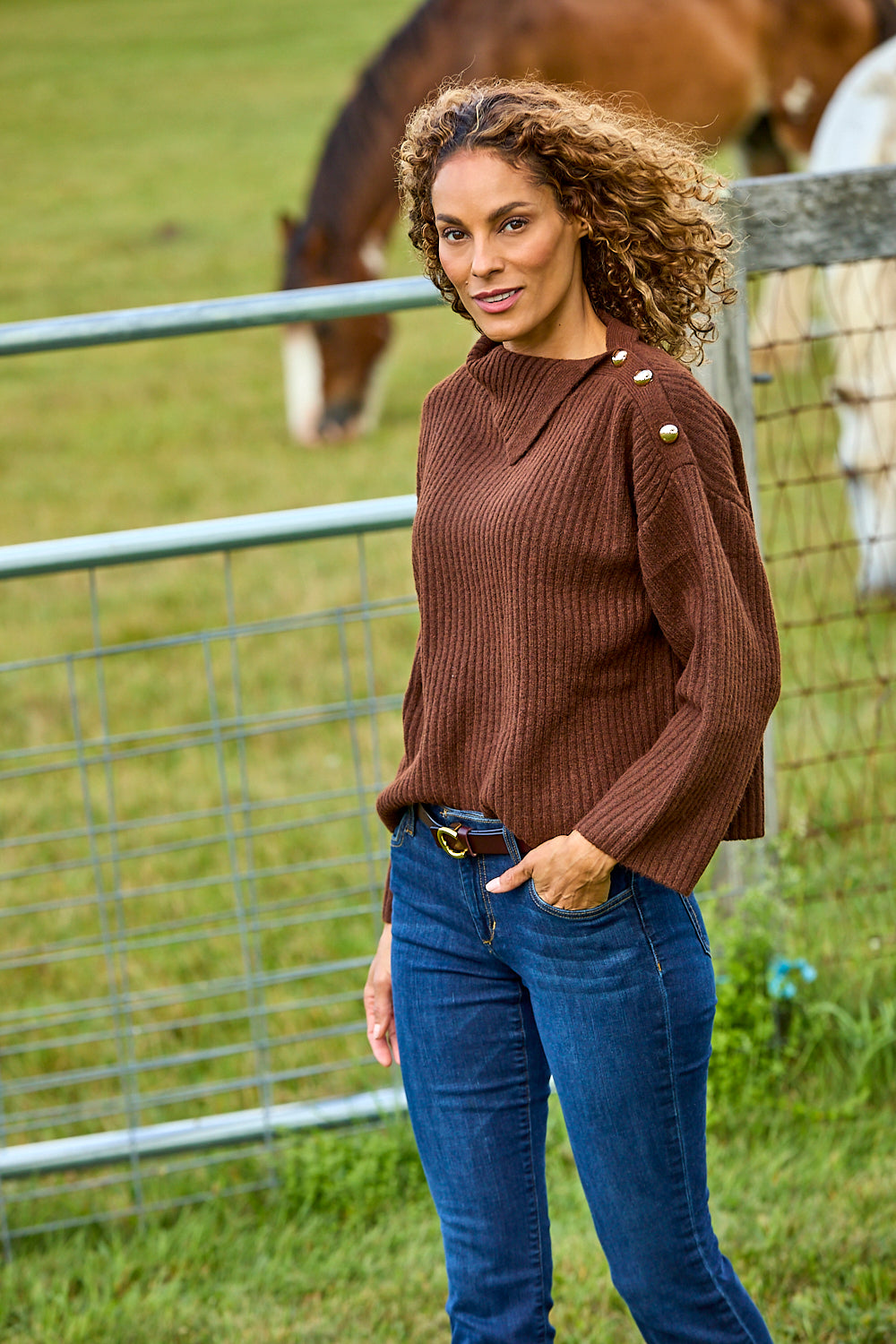 A woman wearing a brown ribbed knit sweater with gold button details on the shoulders and blue jeans, standing in a grassy field with horses behind a wire fence.