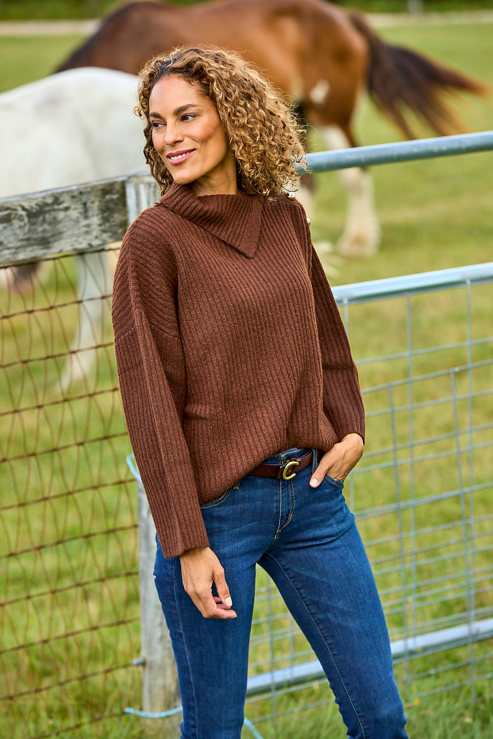 A woman wearing a brown ribbed knit mock neck sweater and blue jeans stands outdoors in front of a wire fence with horses in the background.