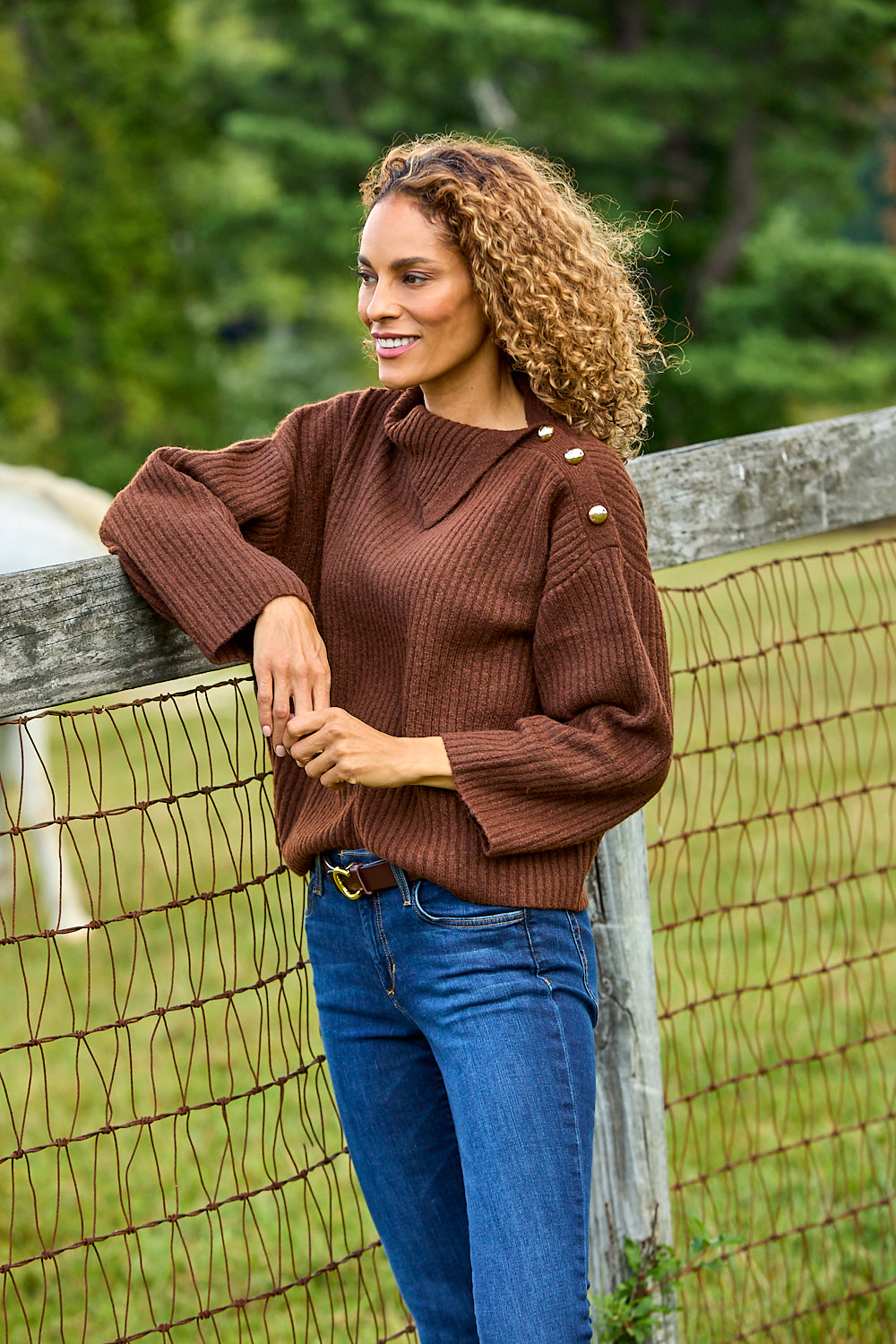 A woman with curly brown hair leans against a rustic wire fence outdoors, wearing a brown ribbed knit sweater with decorative gold buttons on the shoulders and blue jeans.