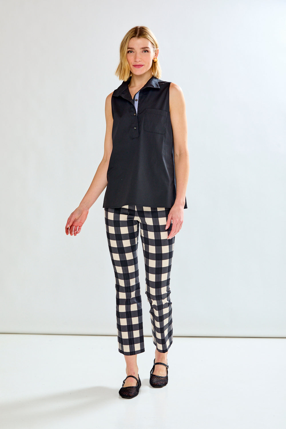 A woman modeling a sleeveless black collared shirt with a button placket, paired with black and white gingham cropped pants and black flats, against a plain white studio background.