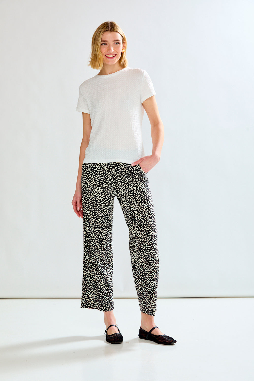 A woman modeling a white short-sleeve knit top and black-and-white patterned cropped trousers with black flats, standing against a clean white studio background.