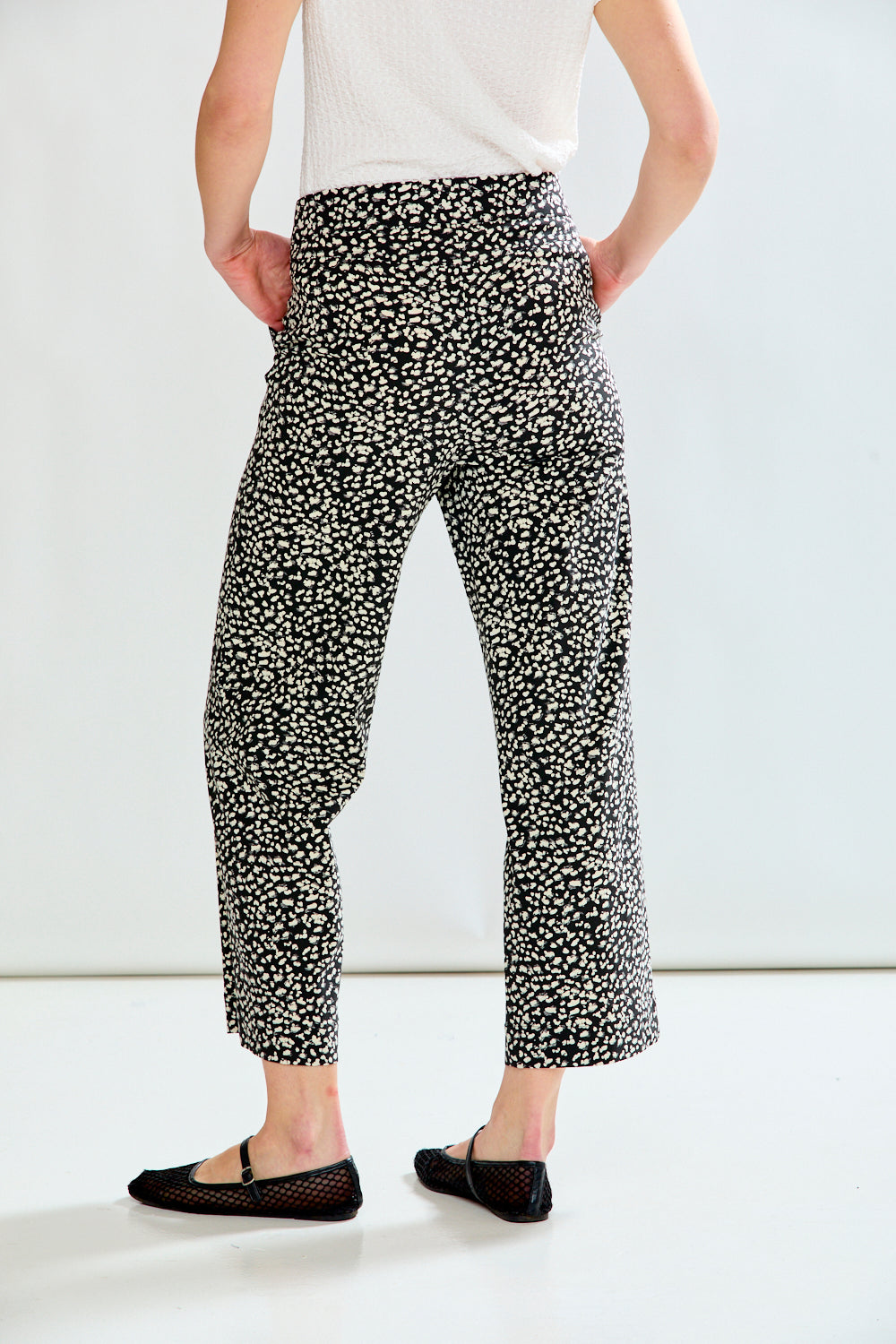 A person shown from the back wearing black cropped pants with a dense white abstract speckled pattern, paired with a white top and black flats, against a plain light studio background.