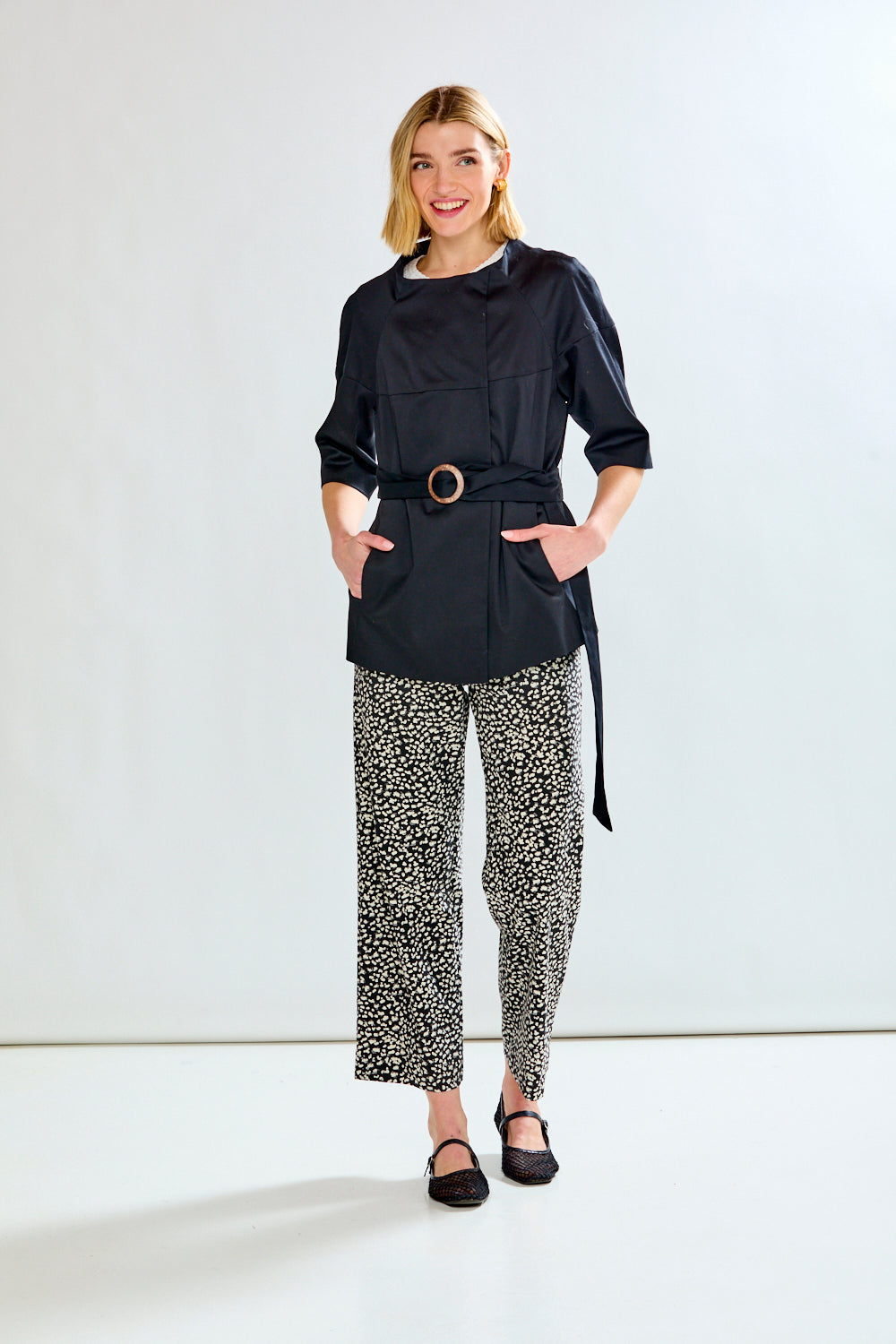 A woman modeling a black belted jacket with a black and white patterned trouser set, standing against a plain white studio background.