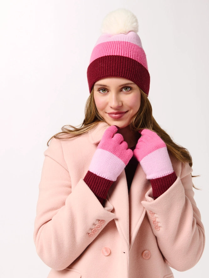 A woman wearing a pink, light pink, and burgundy striped knit beanie with a white pom-pom, matching striped knit gloves, and a light pink coat, posing against a solid white background.