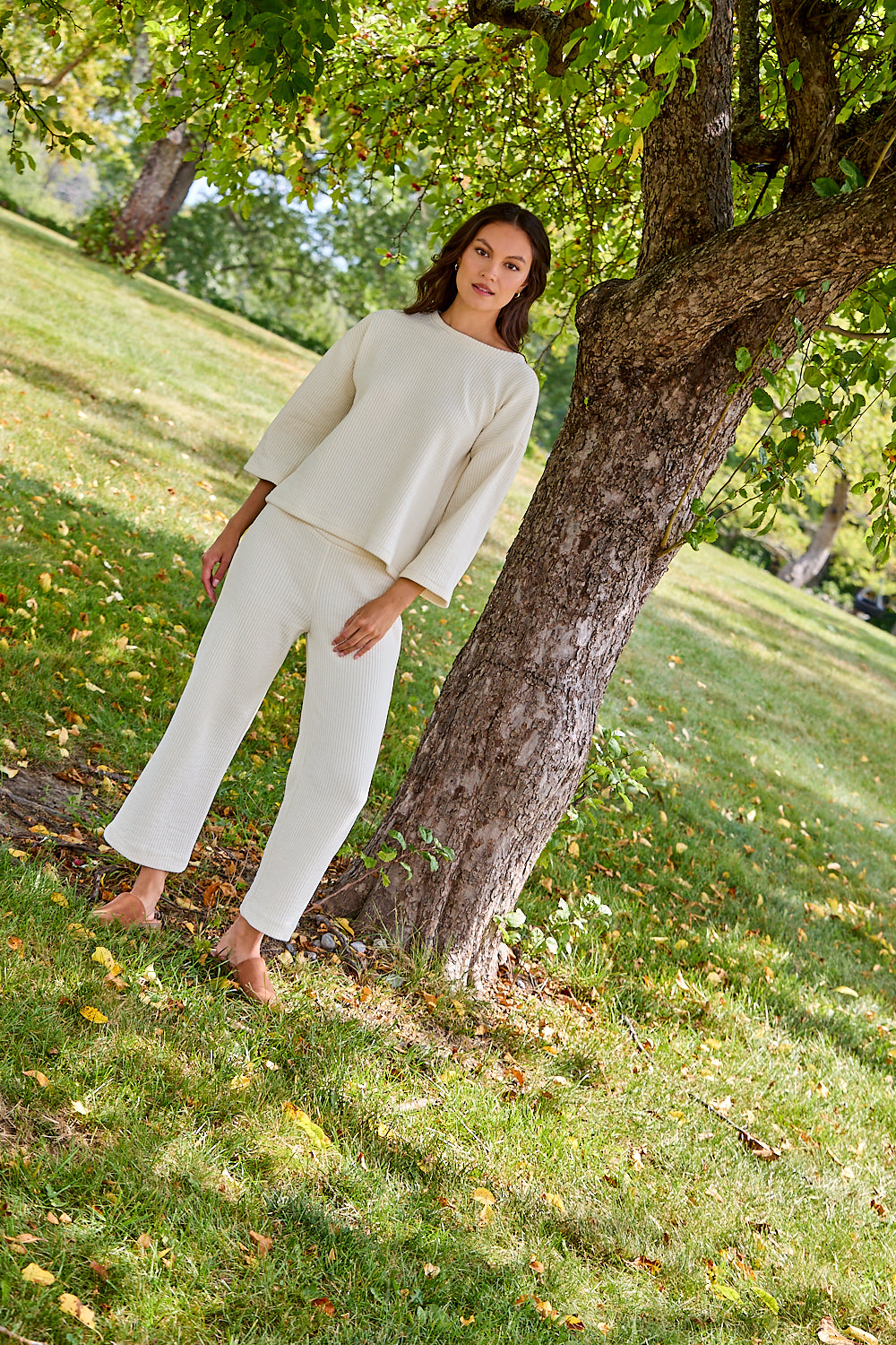 A woman modeling a cream-colored two-piece lounge set consisting of a cropped long-sleeve top and matching wide-leg pants, standing barefoot on a grassy lawn next to a tree in a park.