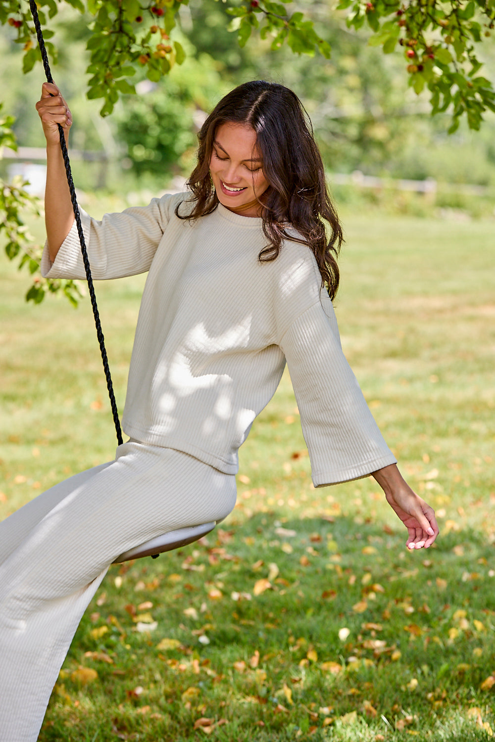A woman wearing a cream-colored textured knit two-piece outfit with a mock neck top and wide flared sleeves, sitting on a swing in a grassy park.