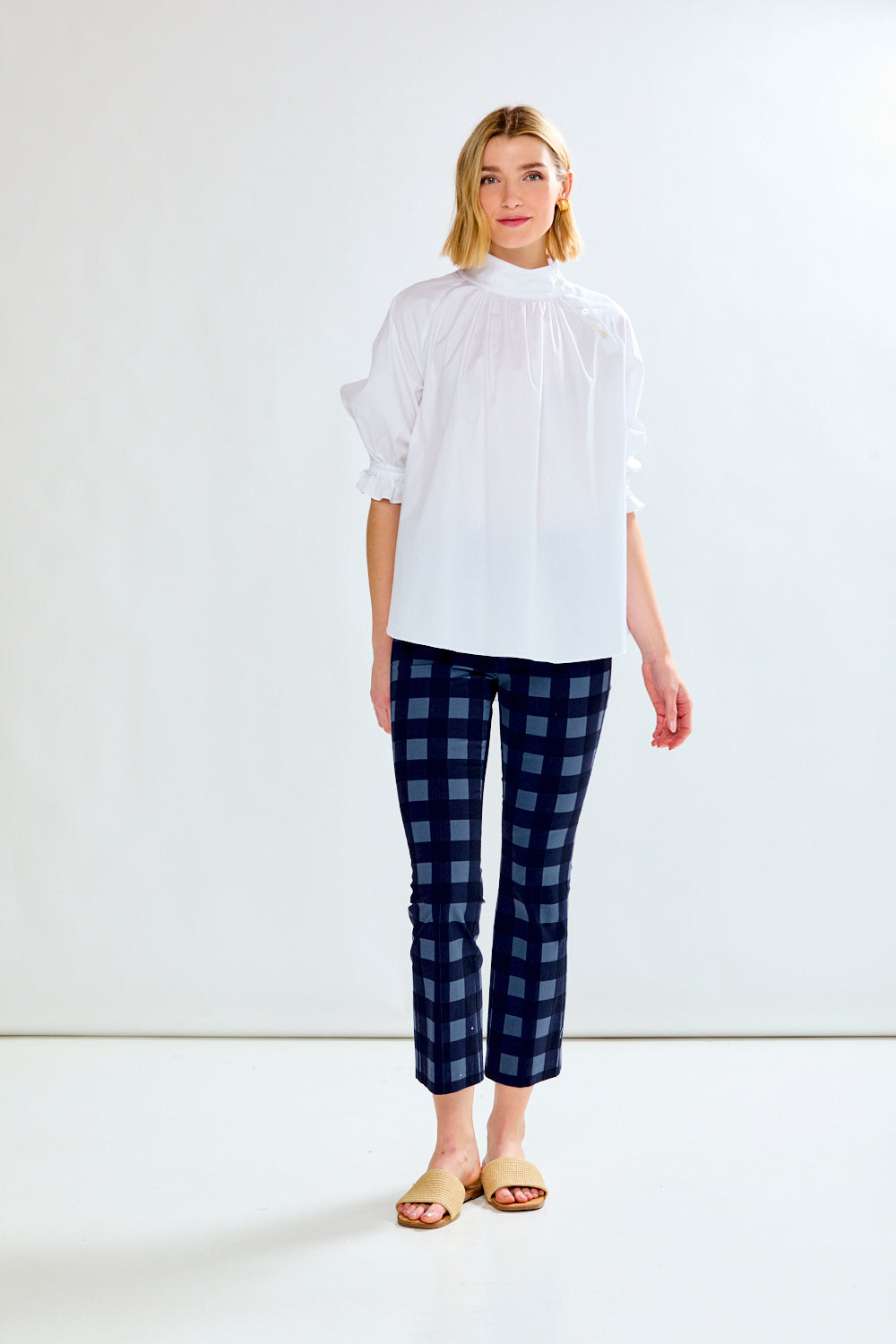 A woman modeling a white high-neck blouse with voluminous sleeves and dark blue plaid cropped pants, standing against a plain white studio background.