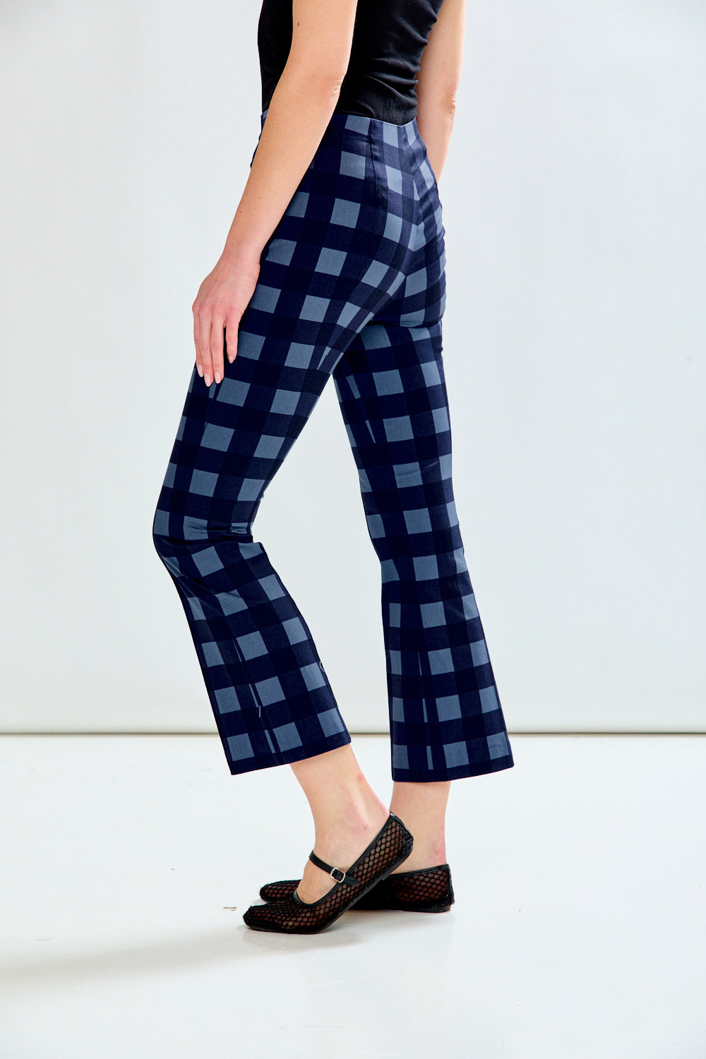 A model is shown from the waist down wearing navy blue gingham cropped pants with a black top and black Mary Jane flats, set against a plain white studio background.