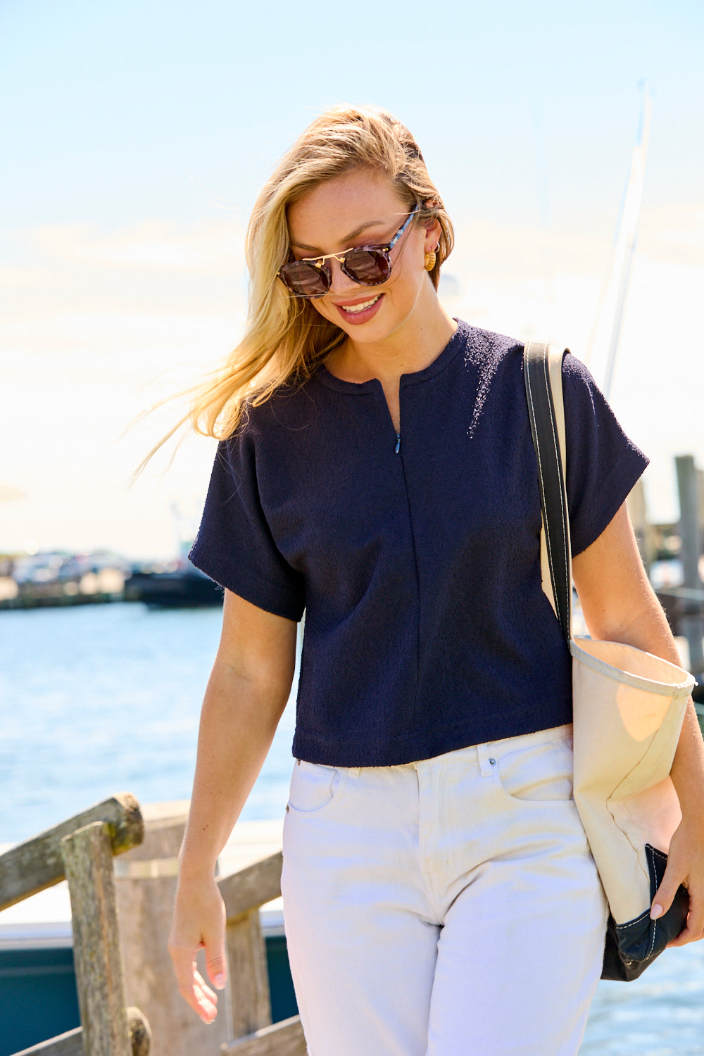 Woman in navy top and white pants standing by a waterfront with sunglasses on