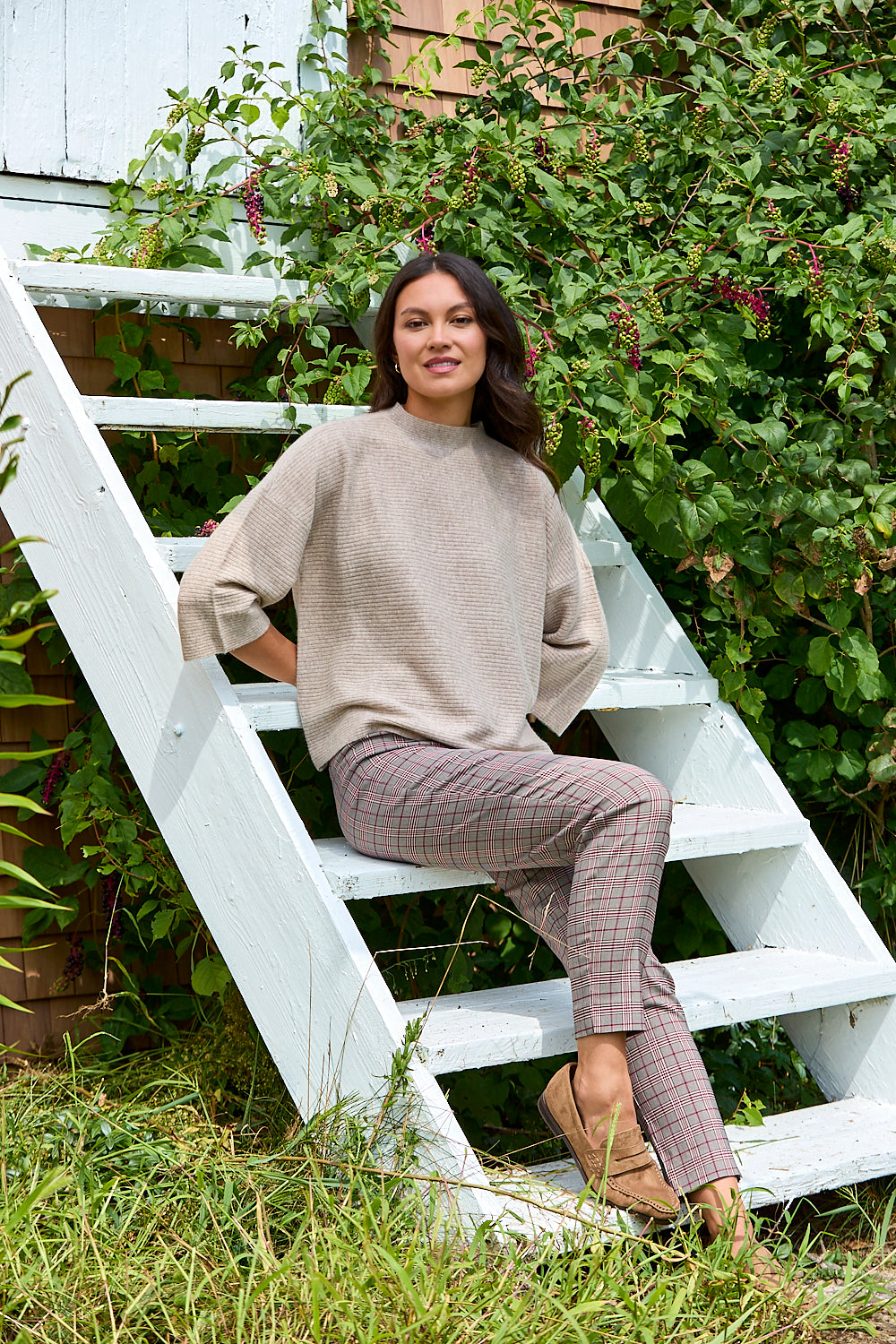 A woman sits on white outdoor stairs surrounded by green plants, wearing a beige knit sweater and plaid trousers.