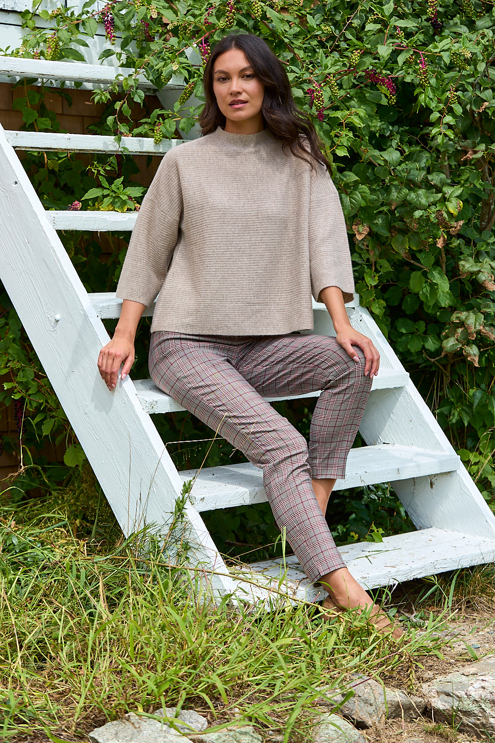 A woman sits on white wooden garden steps outdoors, wearing a beige textured mock-neck top and brown plaid cropped trousers.