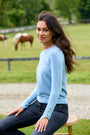 A woman wearing a light blue long-sleeve knit sweater and dark jeans sits on a wooden stool in a rural outdoor setting with horses grazing in the background.