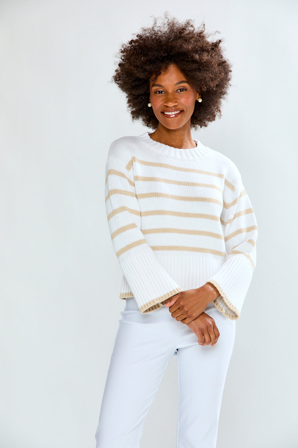 Woman wearing a white and beige striped sweater with white pants on a plain background