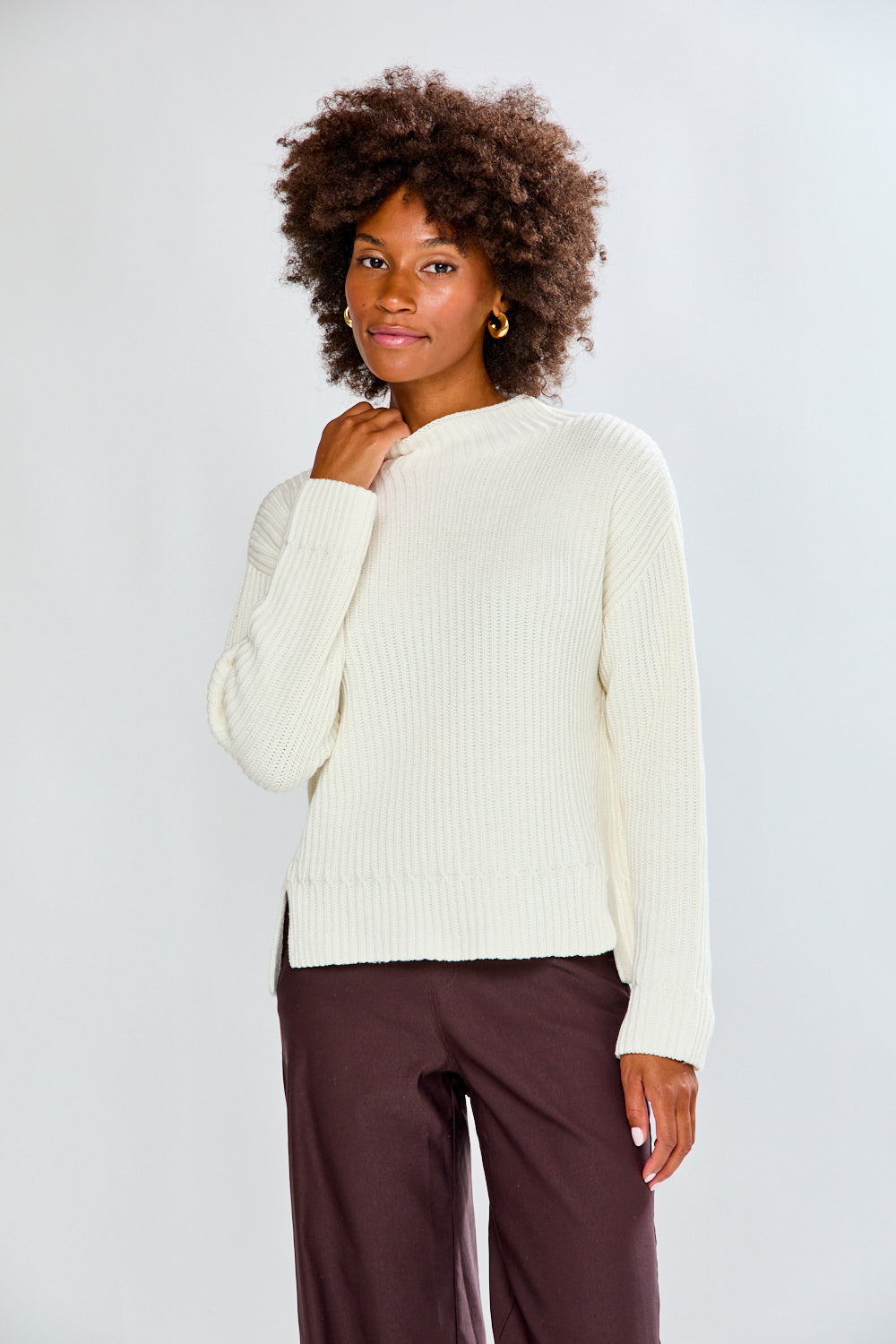Woman wearing a white sweater and brown pants against a plain background