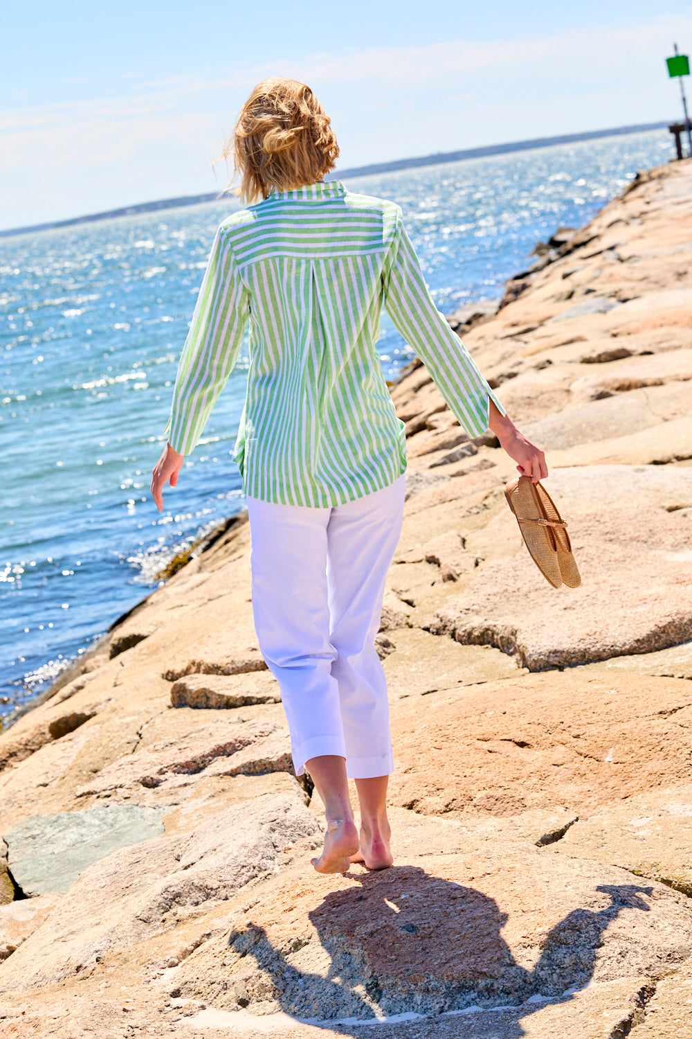 Woman in a striped shirt and white pants walking along a rocky shoreline.