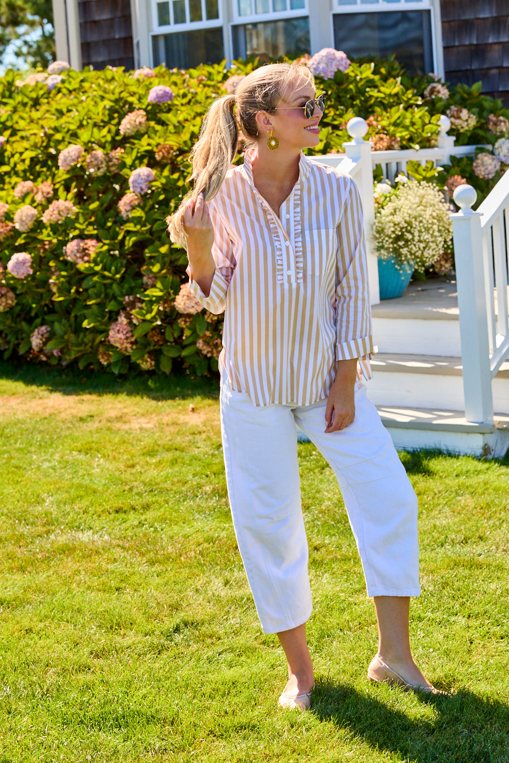 Woman in a striped shirt and white pants standing on grass with flowers and a house in the background