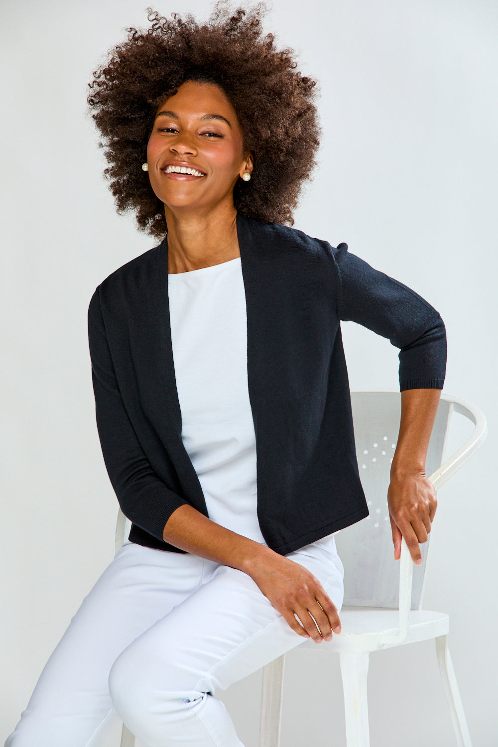 Woman wearing a black blazer over a white shirt, sitting on a white chair against a light gray background.