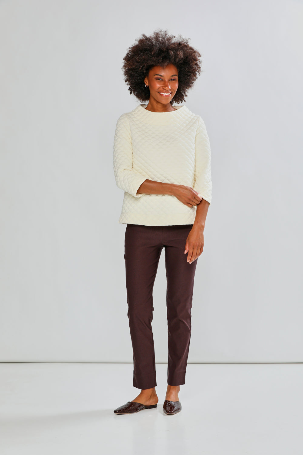 Woman wearing a cream sweater and brown pants against a light gray background