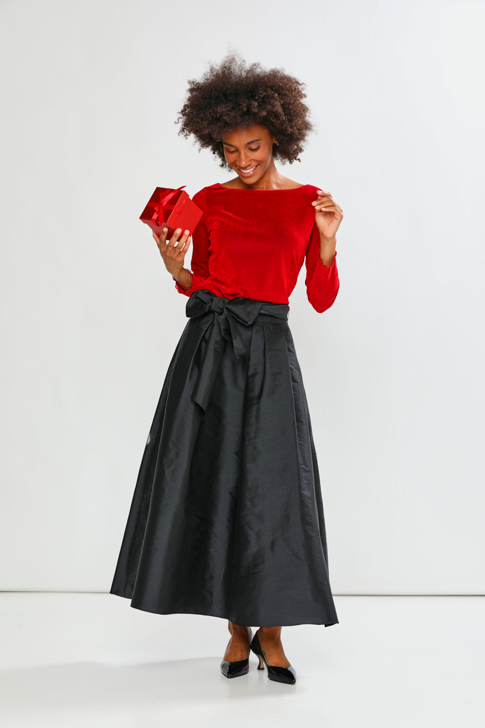 Woman in a red top and black skirt holding a red gift box against a white background