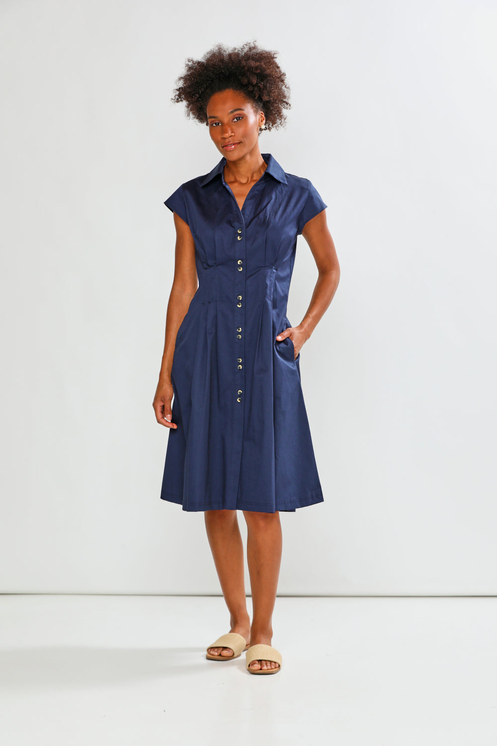 A woman modeling a navy blue short-sleeve midi dress with a button-down front, collar, and side pockets, standing against a plain white studio background.