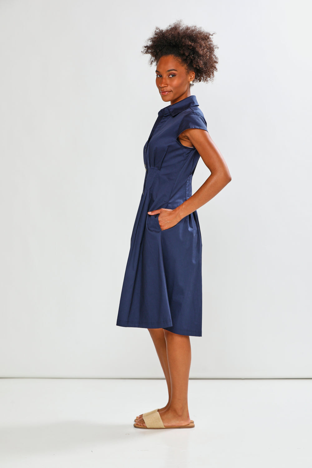 A woman modeling a navy blue short-sleeve midi dress with a high collar and side pockets, paired with beige slide sandals, against a plain white studio background.