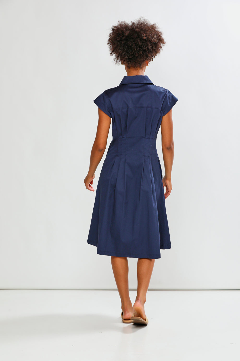 A woman viewed from behind wearing a navy blue short-sleeve collared dress with a gathered waist and flared skirt, standing against a plain light gray studio background.
