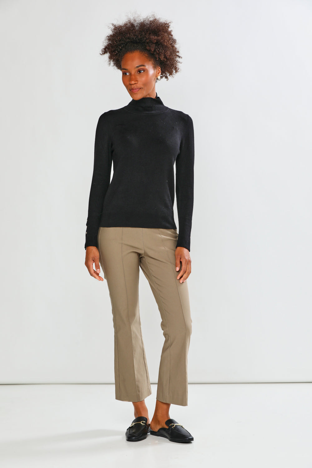 A woman modeling a black long-sleeve mock neck top and mushroom cropped trousers with black loafers, standing against a plain white studio background.