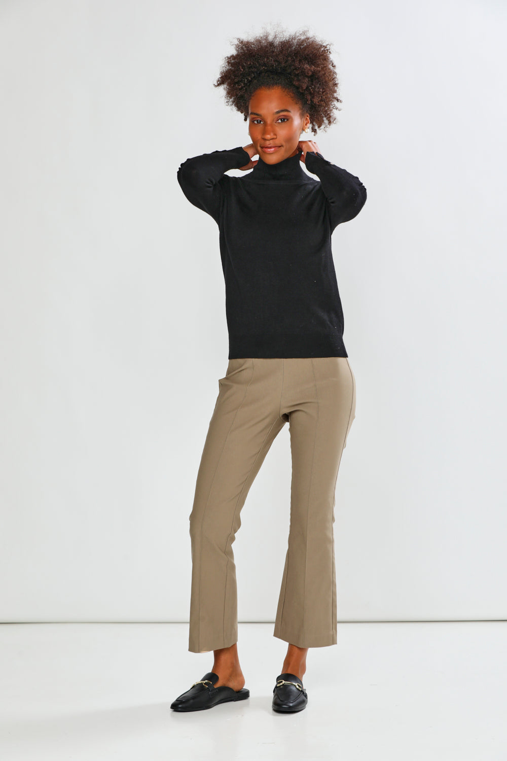 A woman modeling a black long-sleeve mock neck top and mushroom cropped trousers with black loafers, standing against a plain white studio background.