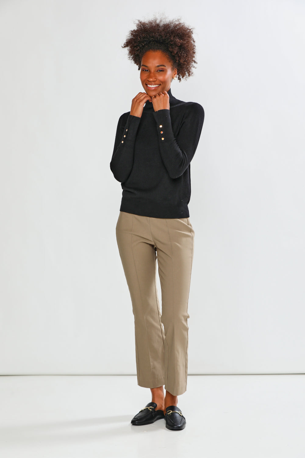 A woman modeling a black long-sleeve mock neck top and mushroom cropped trousers with black loafers, standing against a plain white studio background.