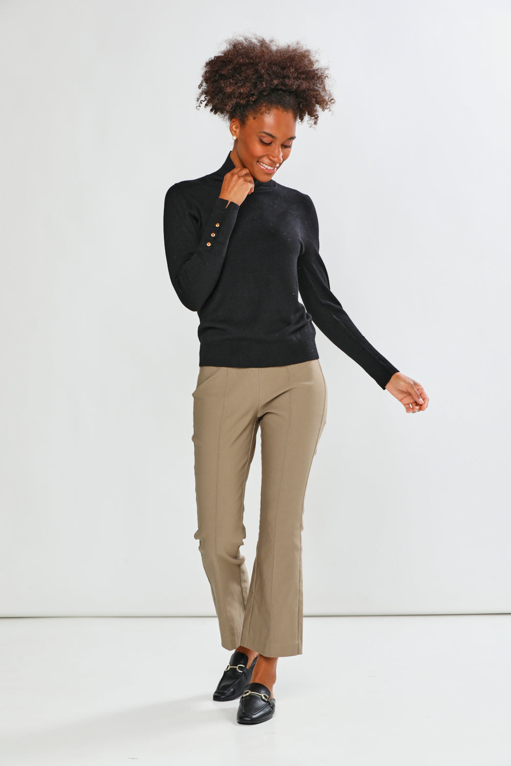 A woman modeling a black long-sleeve mock neck top and mushroom cropped trousers with black loafers, standing against a plain white studio background.