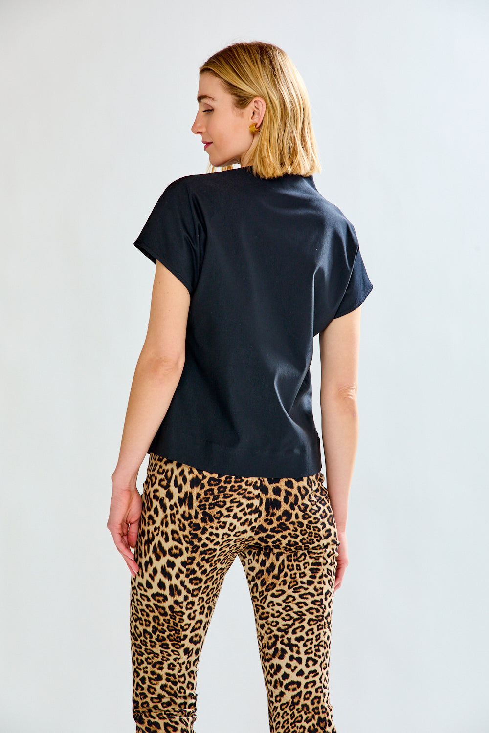 A woman shown from the back wearing a black short-sleeve top and leopard-print pants against a light gray studio background.