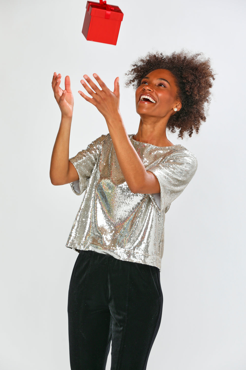 Woman in a silver sequin top catching a red gift box against a white background