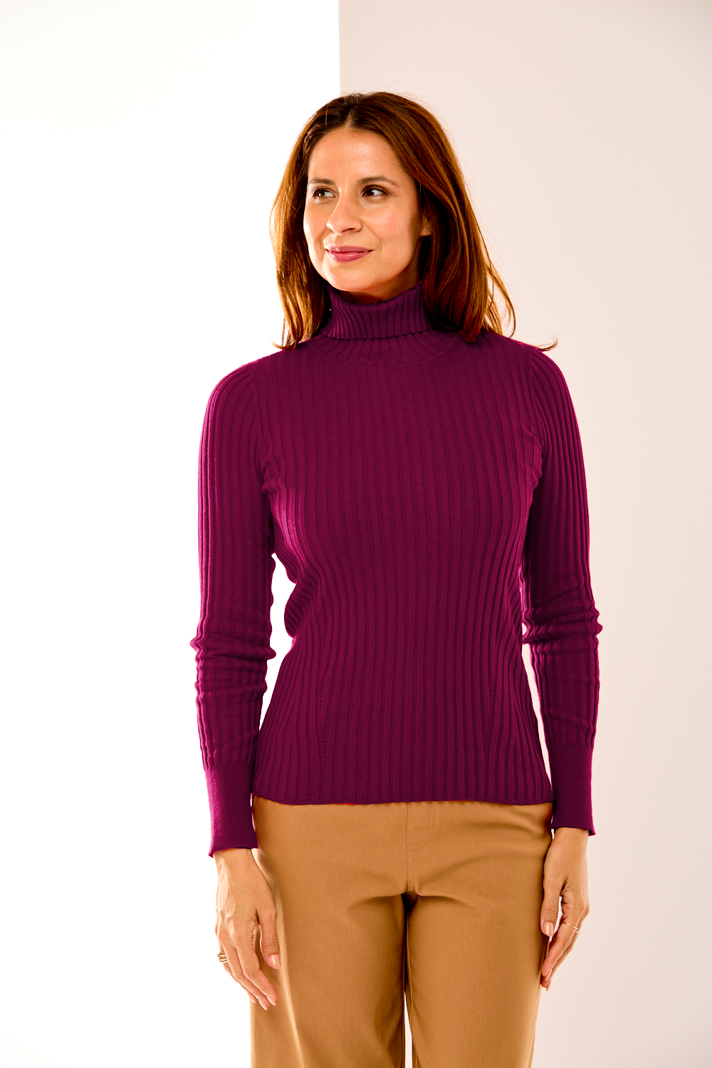 Woman wearing a purple turtleneck sweater and beige pants against a white background