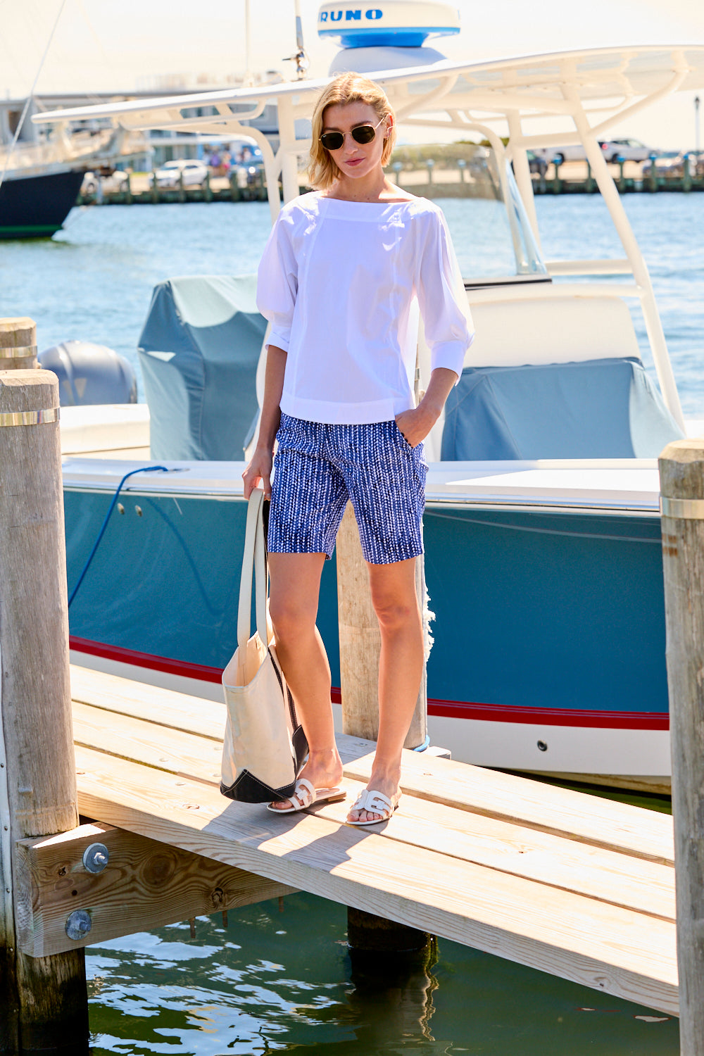 Woman standing on a dock by a boat, wearing a white shirt and patterned shorts.