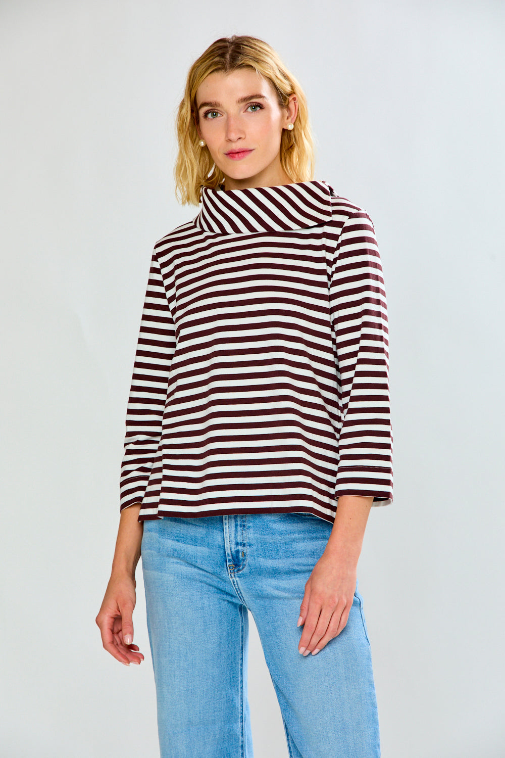 A woman modeling a brown and white horizontally striped knit top with a wide mock neck and three-quarter sleeves, paired with light blue jeans, against a light gray studio background.